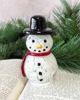 Snowman figurine with a black top hat and red scarf on an open book with greenery in the background
