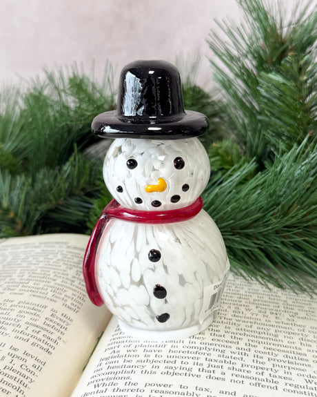 Snowman figurine with a black top hat and red scarf on an open book with greenery in the background