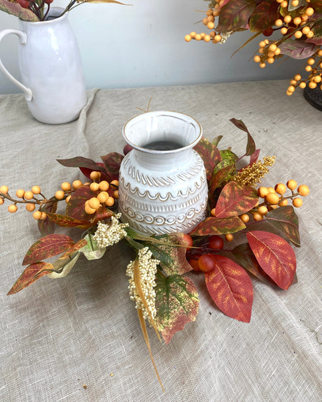 Decorative arrangement with a white vase and autumn-themed leaves on a textured surface.